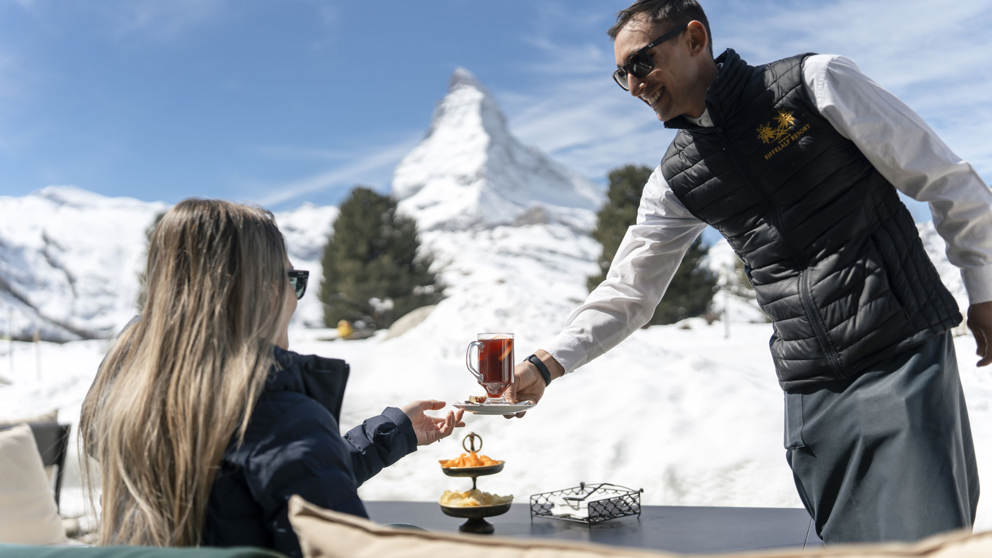 Serveur apportant un cocktail sur la terrasse du Bar 2222m avec vue sur le Matterhorn au Riffelalp Resort 2222m à Zermatt, hôtel de luxe 5 étoiles dans les Alpes suisses proposant une expérience après-ski haut de gamme dans un cadre alpin exclusif.