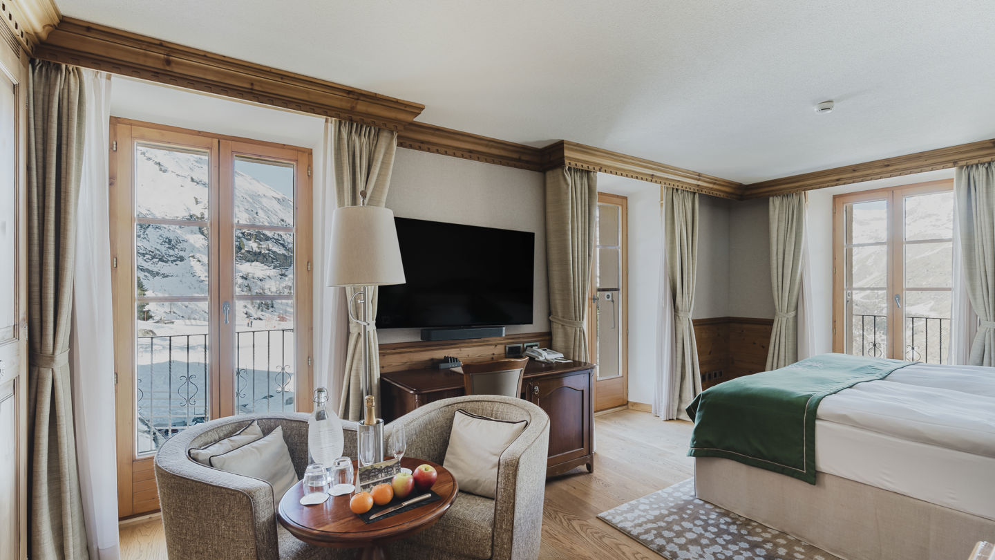 Vue d’ensemble du coin salon, du lit et des fenêtres sur les montagnes enneigées de la Chambre NATUR Grandlit en hiver au Riffelalp Resort 2222m à Zermatt, hébergement chaleureux avec petit-déjeuner et accès au Spa 2222m inclus.